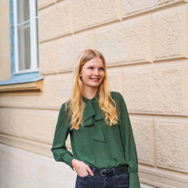 Frau mit Bluse steht neben einer Mauer und schaut an der Kamera vorbei