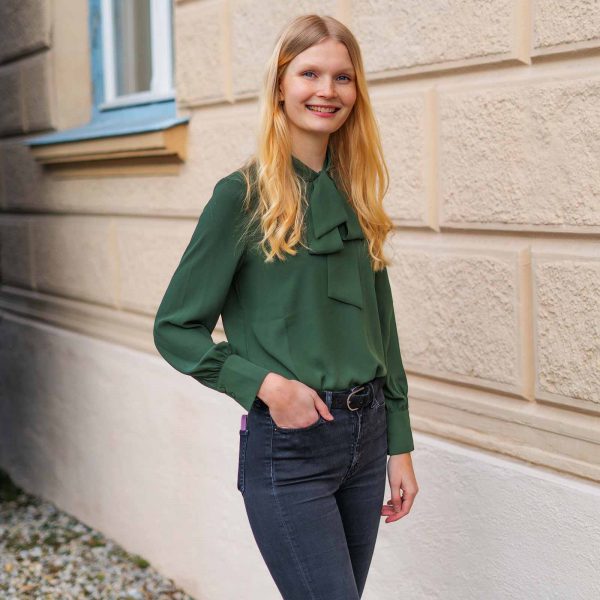 Frau mit Bluse steht neben einer Mauer und schaut in die Kamera