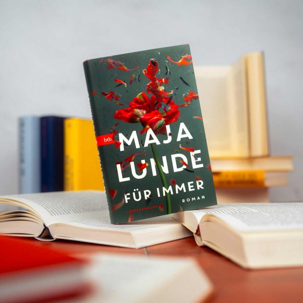 Das Buch Für Immer auf zwei aufgeklappten Büchern, dahinter sind weitere Offene oder bunte Bücher zu ssehen